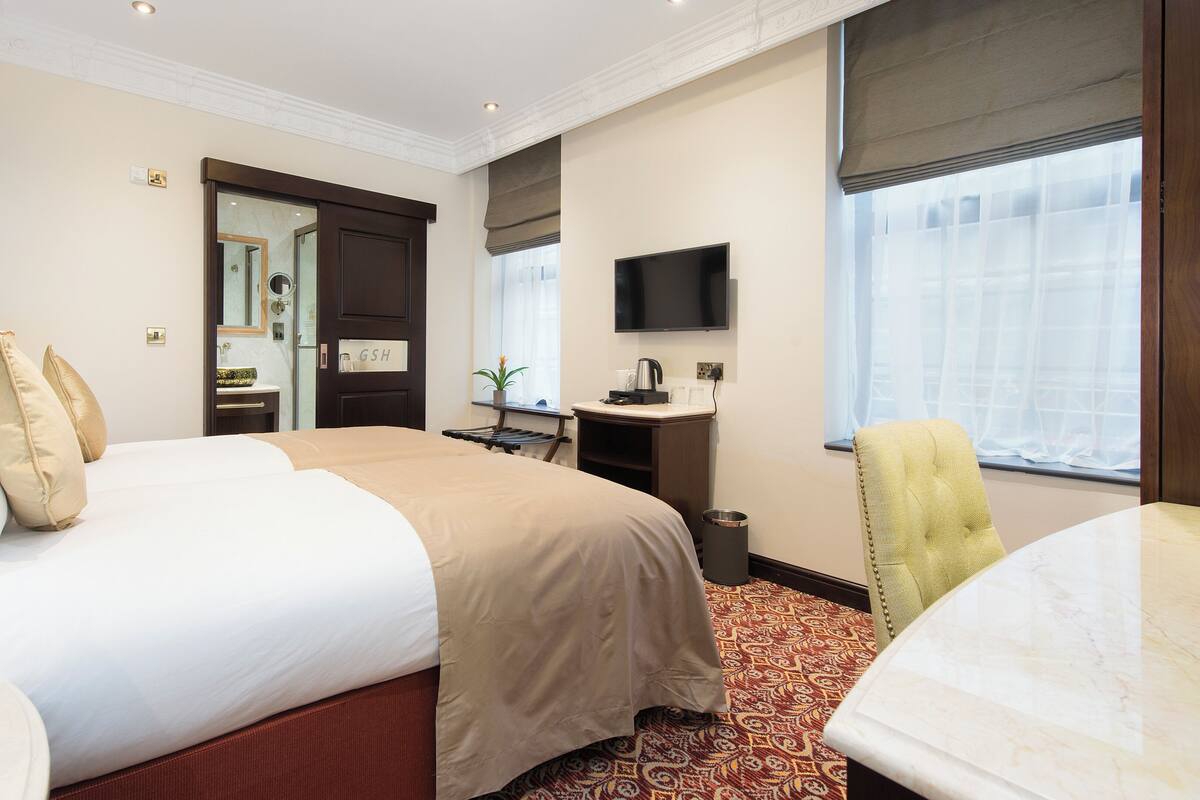 A hotel room with a large bed, a desk with a flat-screen TV, a small window with curtains, and a patterned carpet.
