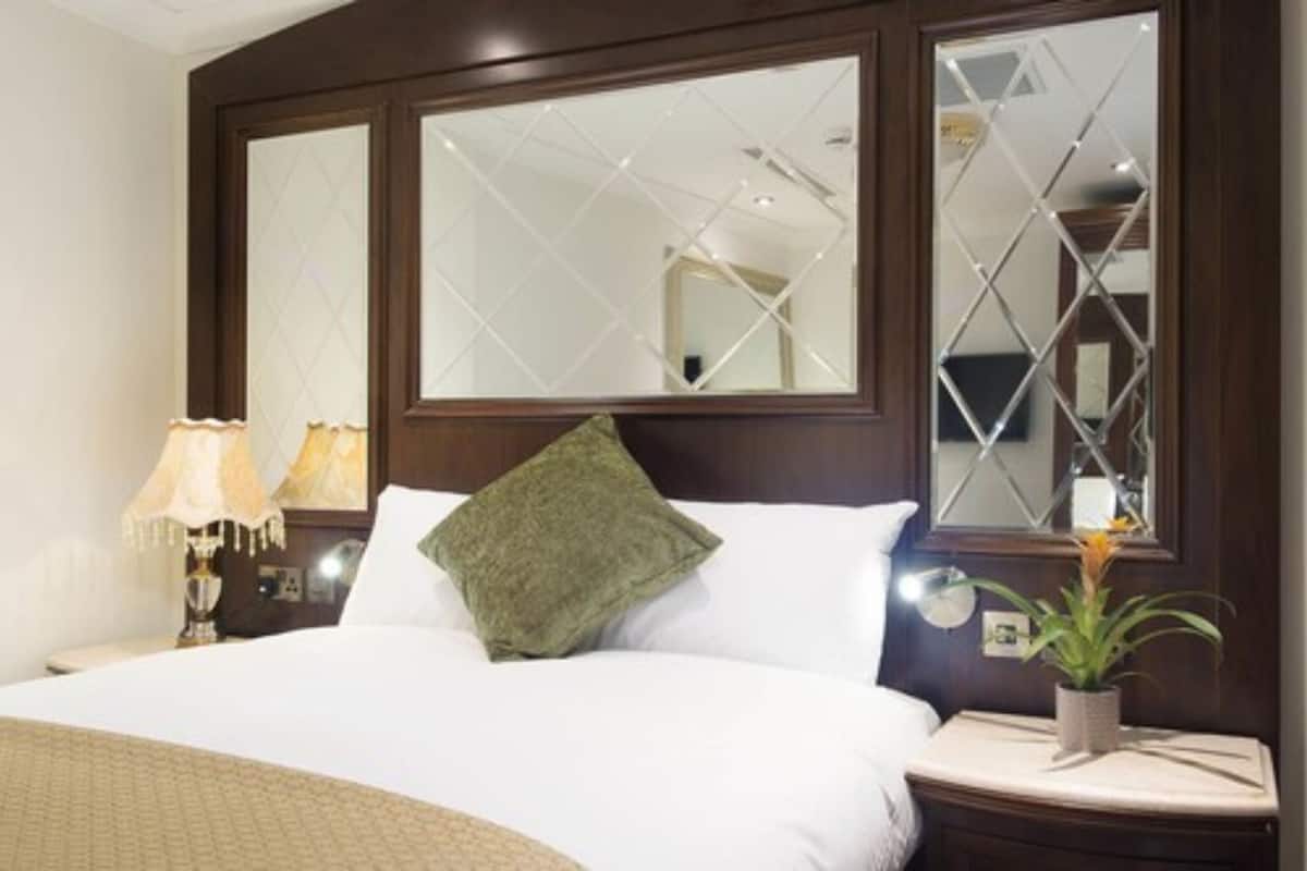 A hotel room with a large bed, two bedside lamps, a nightstand with a plant, and a mirror with a geometric pattern.