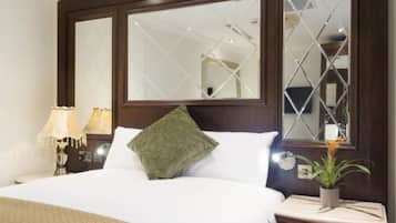 Quarto de hotel com uma cama grande, duas luminárias de cabeceira, uma mesinha de cabeceira com uma planta e um espelho com padrão geométrico.