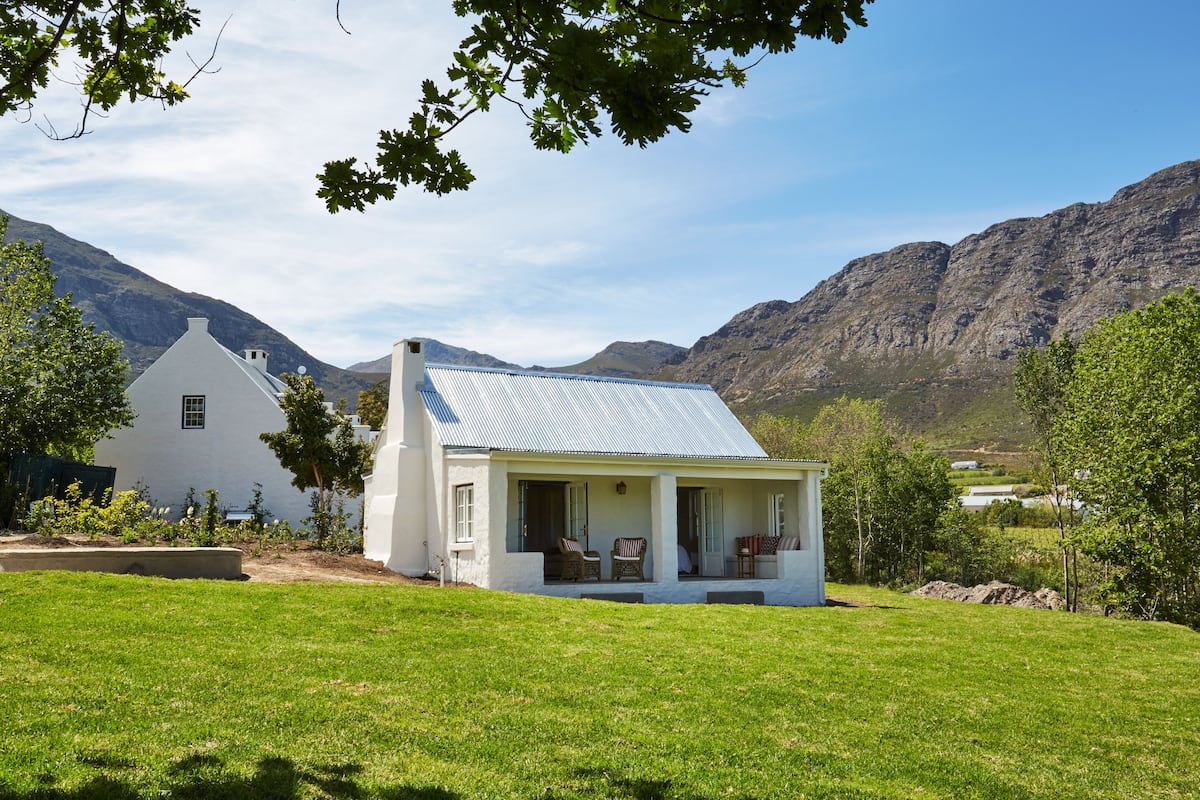 Une maison blanche avec un toit en métal, entourée d’un champ verdoyant et bordée de montagnes en arrière-plan.