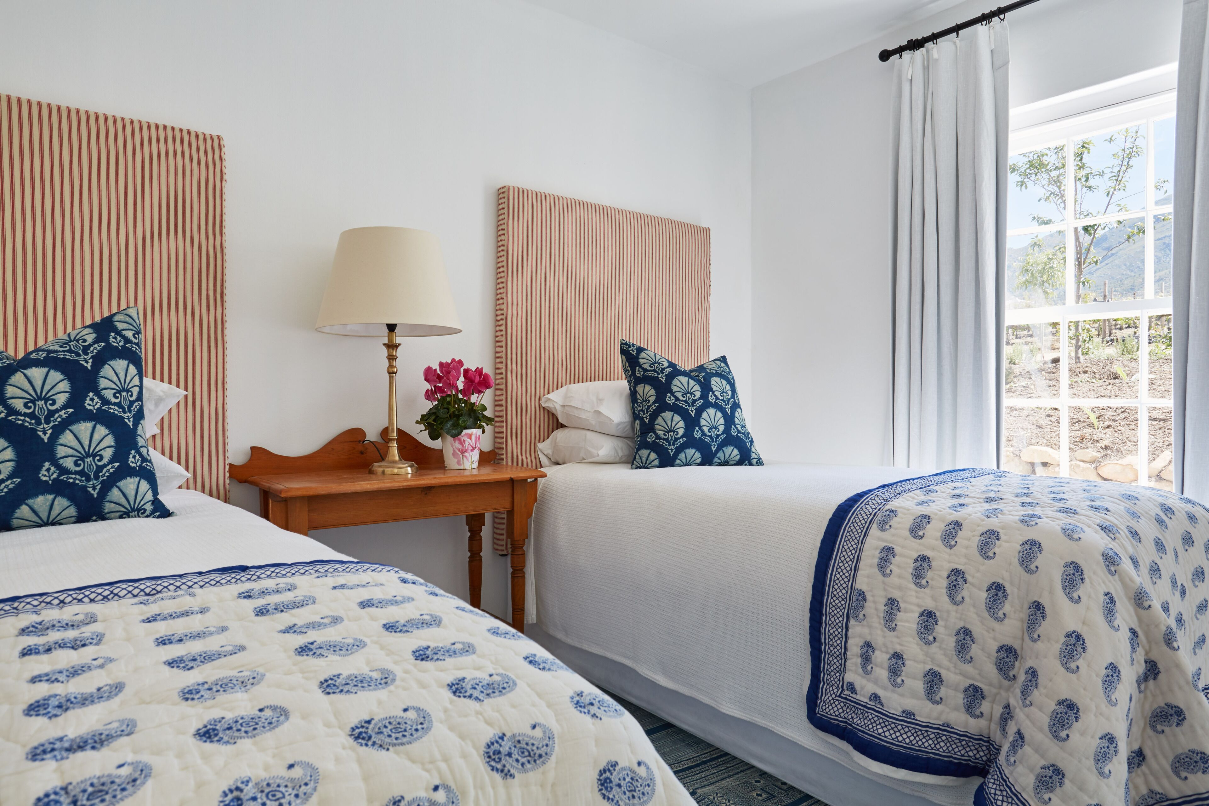 Habitación de hotel con dos camas, una mesita de noche con lámpara y flores, y una ventana con cortinas.
