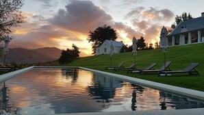 A swimming pool with lounge chairs and umbrellas, a white building with a sloped roof, and a sunset in the background.