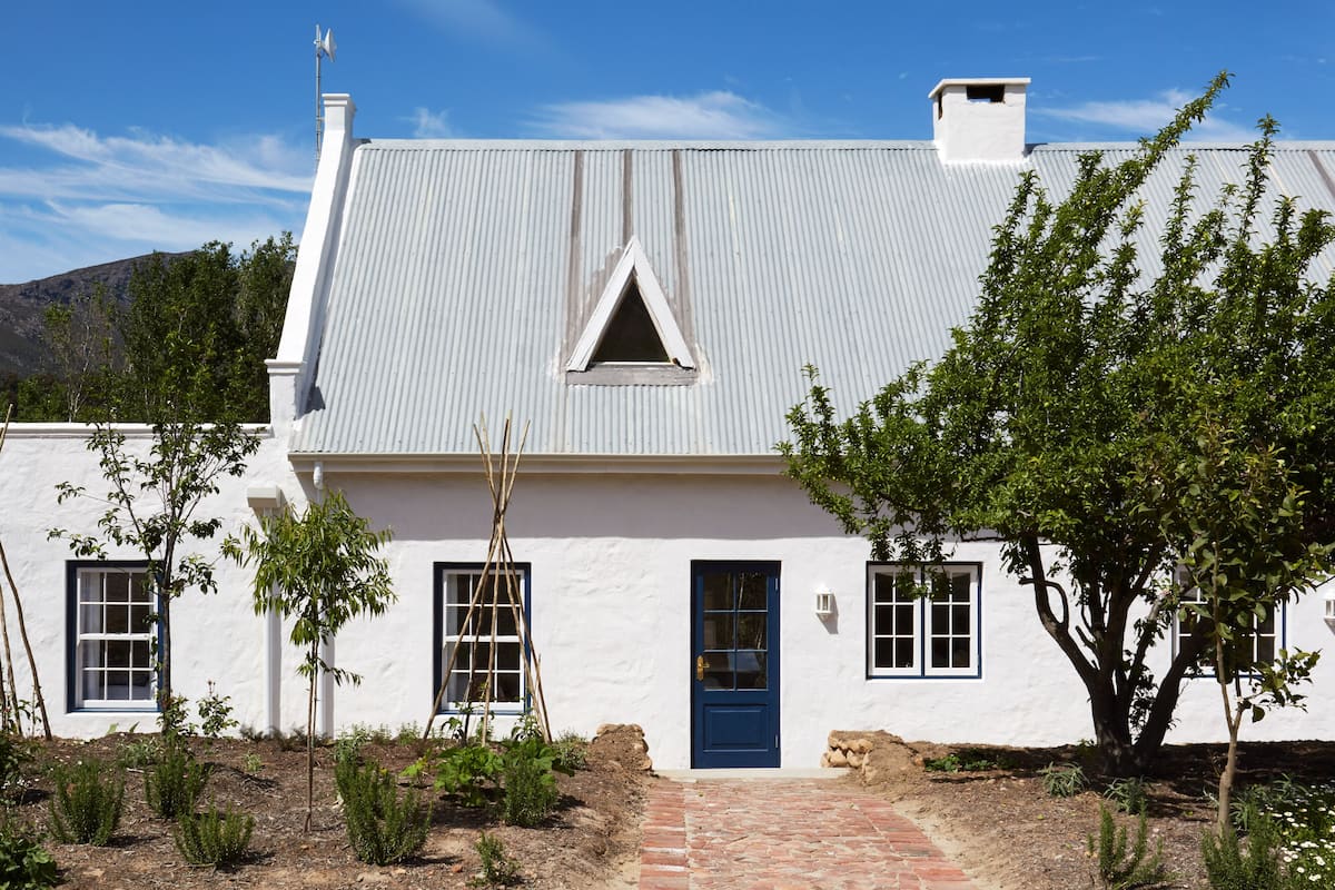Un bâtiment blanc avec une porte et des fenêtres bleues, entouré d’arbres et bordé d’un chemin en briques.