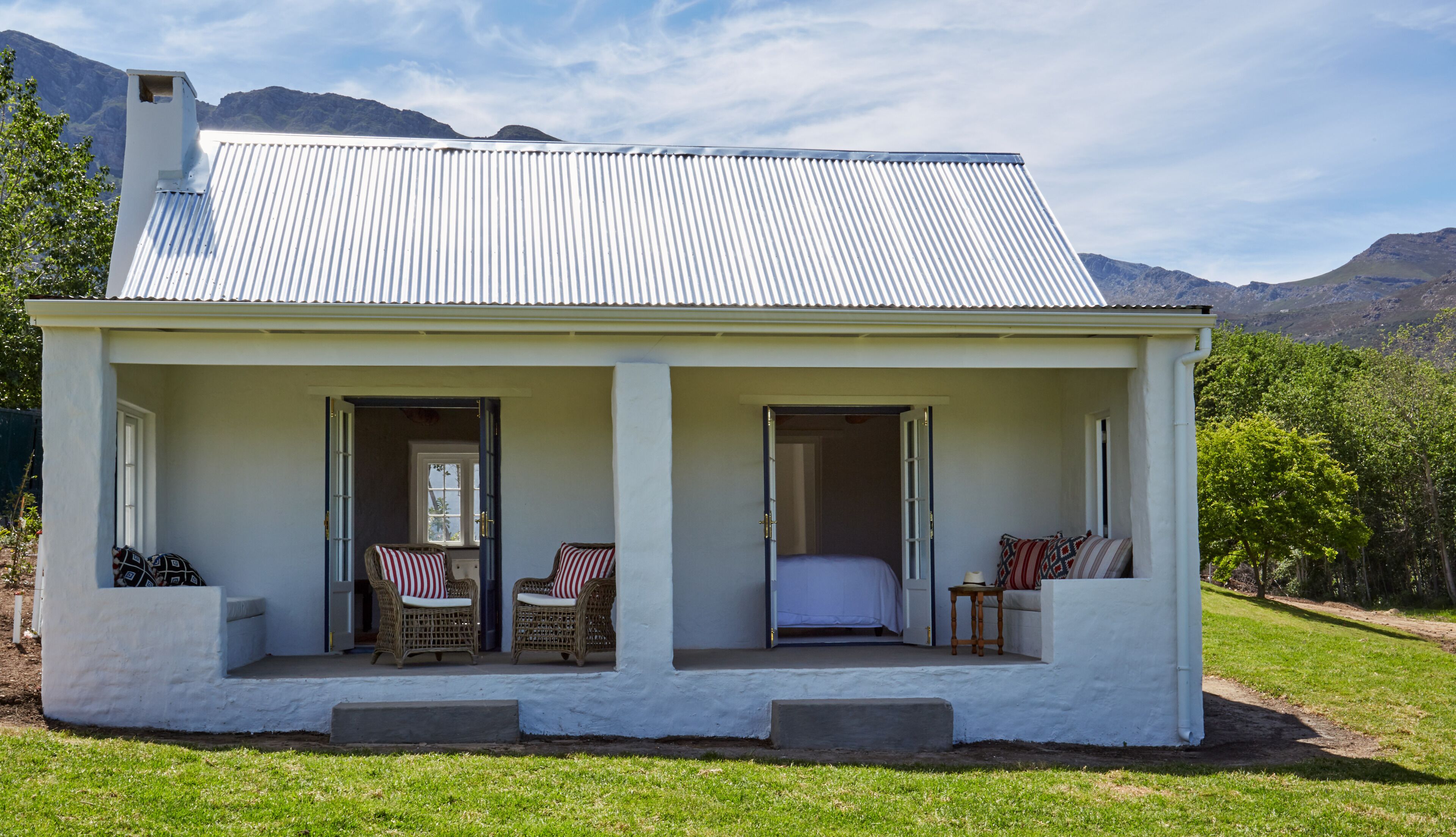 A white cottage with a corrugated metal roof, two open doors leading to a porch with wicker furniture and a bed, set against a mountainous backdrop.