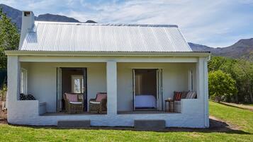 A white cottage with a corrugated metal roof, two open doors leading to a porch with wicker furniture and a bed, set against a mountainous backdrop.