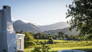 A white building with a window, surrounded by a garden and a pool, with mountains in the background.