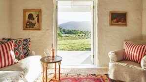 A room with a white couch, a striped armchair, a small table with a wine glass, and a view of a vineyard through an open door.