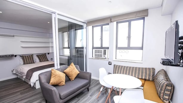 A modern hotel room with a bed, a gray sofa, a white dining table, and a small seating area with cushions. - 106 on Adderley Accommodation (Cape Town)