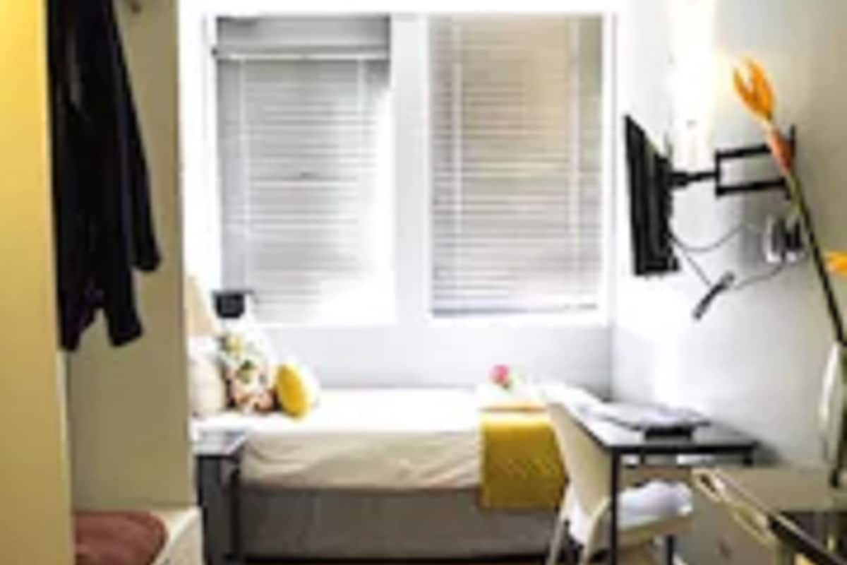 Une petite pièce, de taille modeste, comprenant un lit, un bureau et une chaise. On y trouve une fenêtre avec des stores, un téléviseur fixé au mur et une petite étagère sur laquelle sont posés quelques objets.