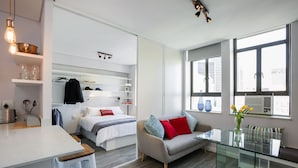 A modern, well-lit room with a glass dining table, a sofa with cushions, a bed with a headboard, and a kitchen area with a countertop and stools. - 106 on Adderley Accommodation (Cape Town)