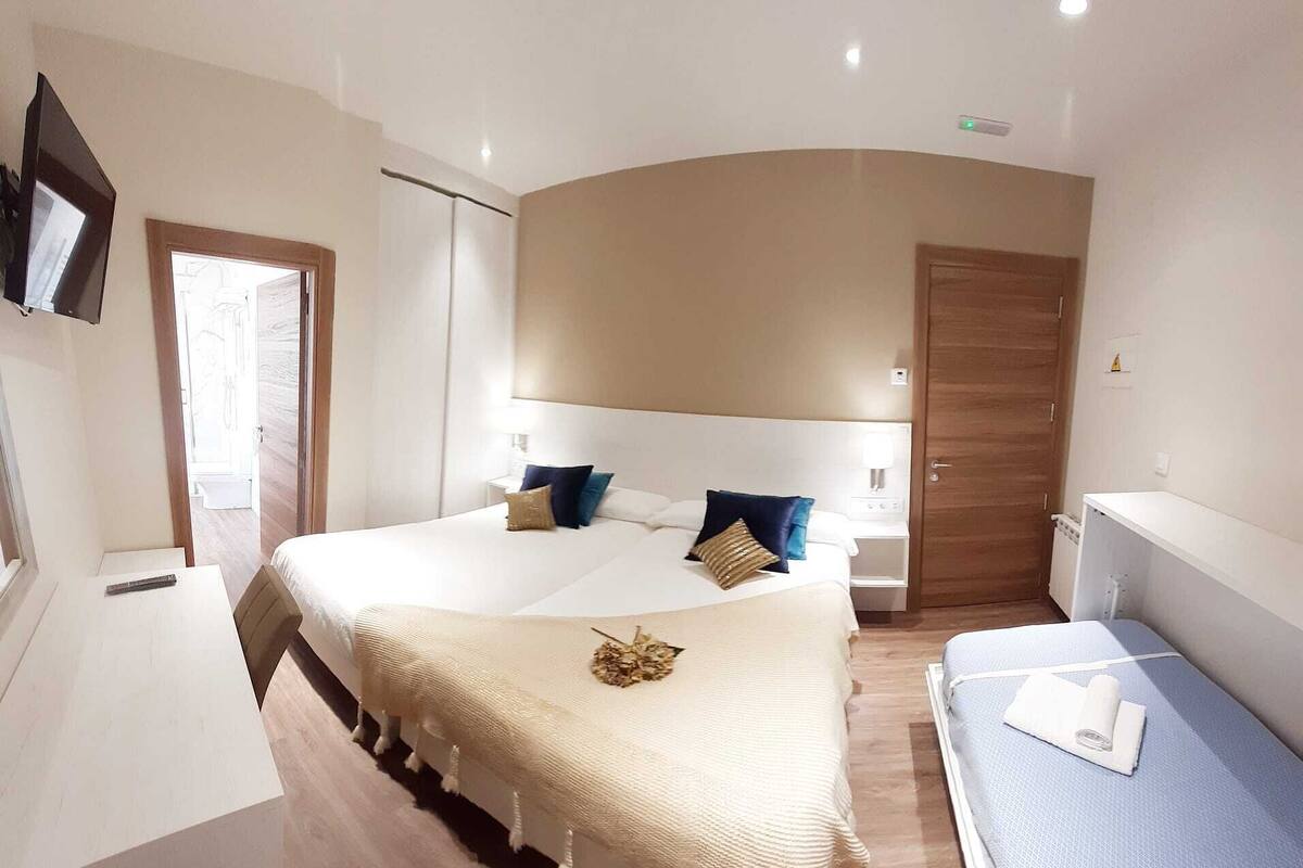 Ein Schlafzimmer mit einem großen Bett, einem kleineren Bett, einem Schreibtisch und einem an der Wand befestigten Fernseher.