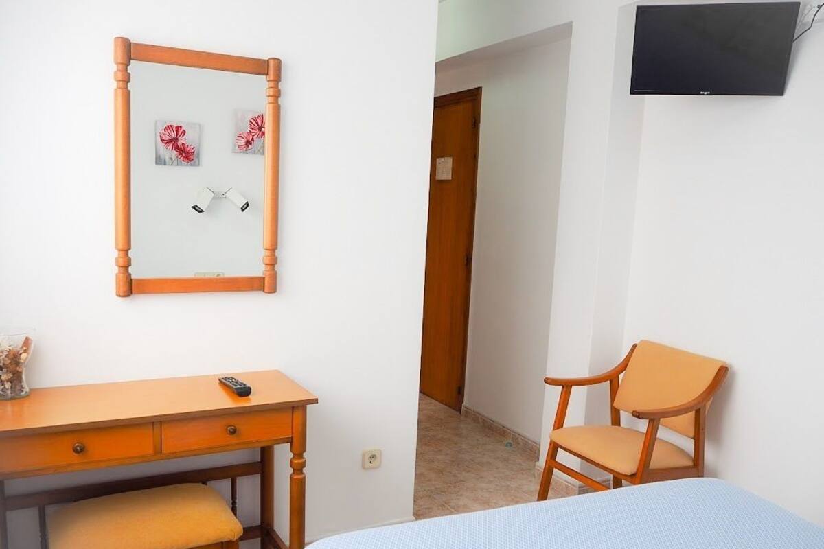 Ein Schlafzimmer mit einem Bett, einem hölzernen Schreibtisch mit Stuhl, einem weiteren Holzstuhl, einem Fernseher und einem Spiegel mit Kunstwerken.