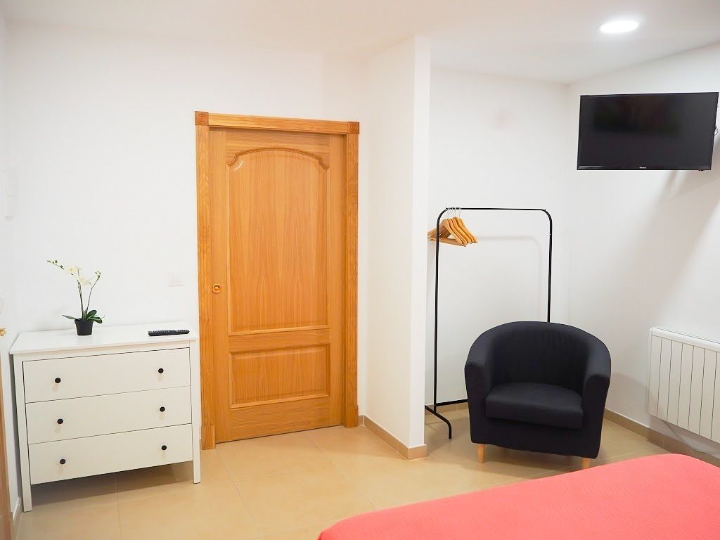 A bedroom with a wooden door, a white dresser, a black chair, a pink bed, and a mounted TV.