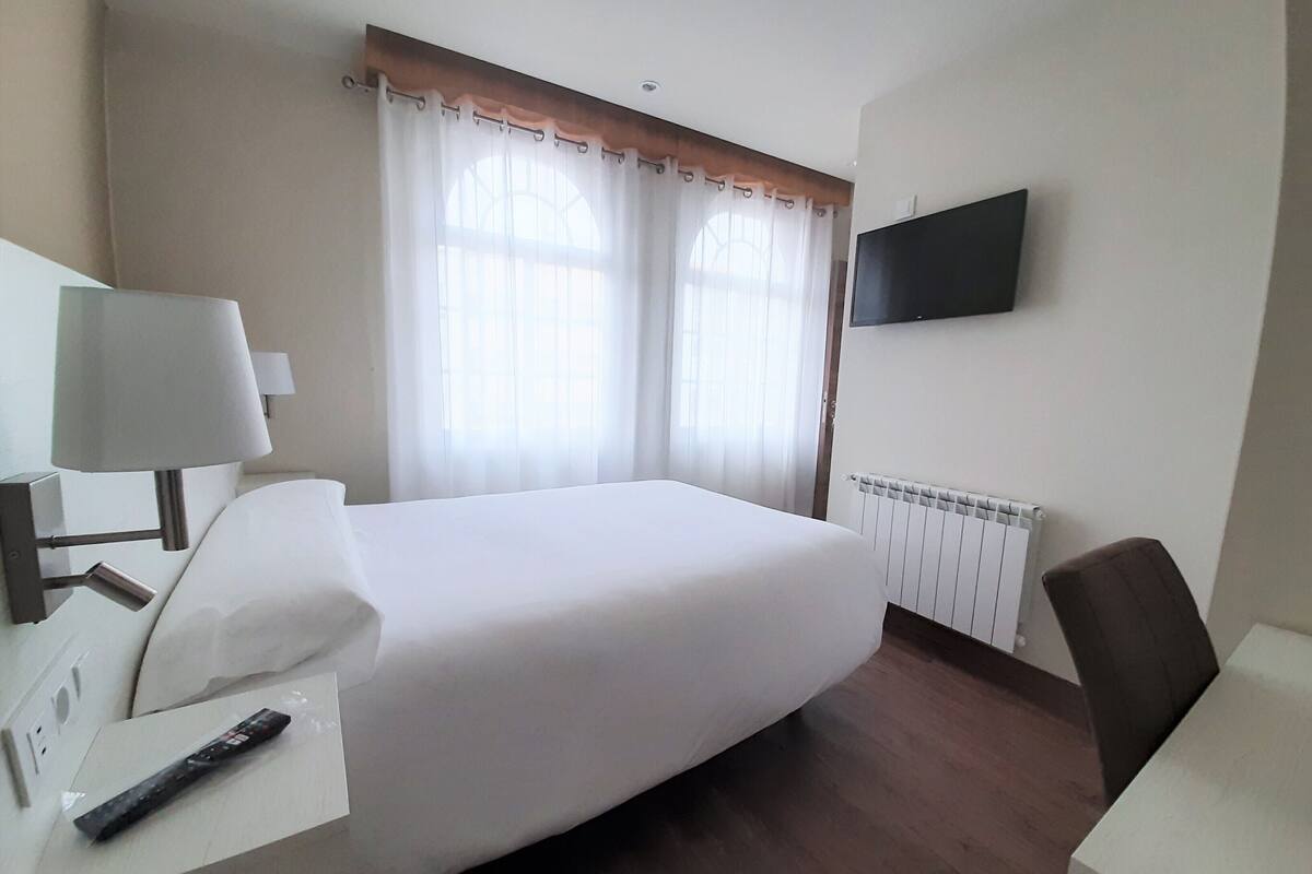 Ein Hotelzimmer mit einem Bett, einem Schreibtisch mit Lampe, einem Stuhl, einem an der Wand befestigten Fernseher und einem Fenster mit Vorhängen.