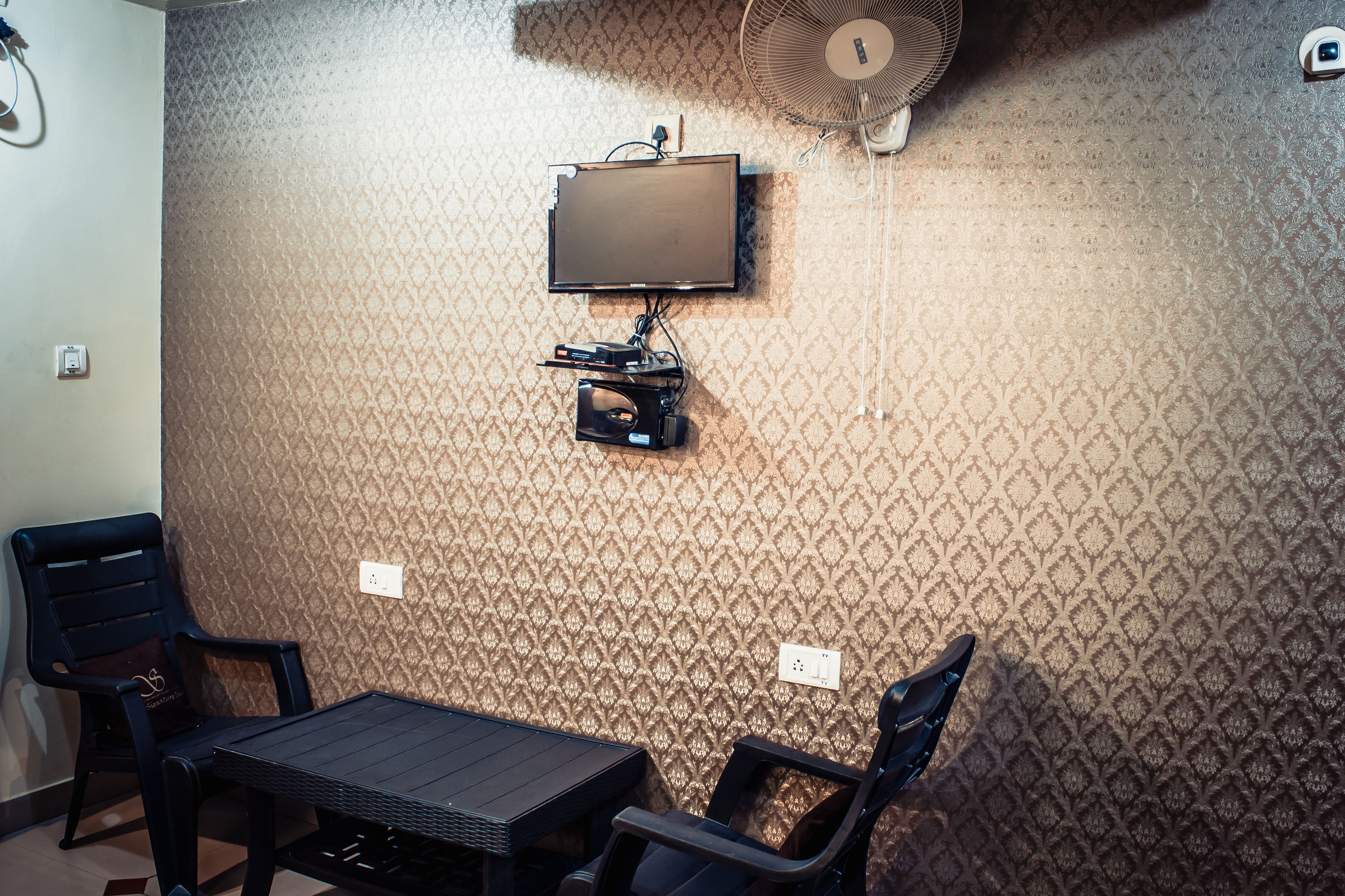 A room with a patterned wallpaper, a mounted television, a table, two chairs, and a standing fan.