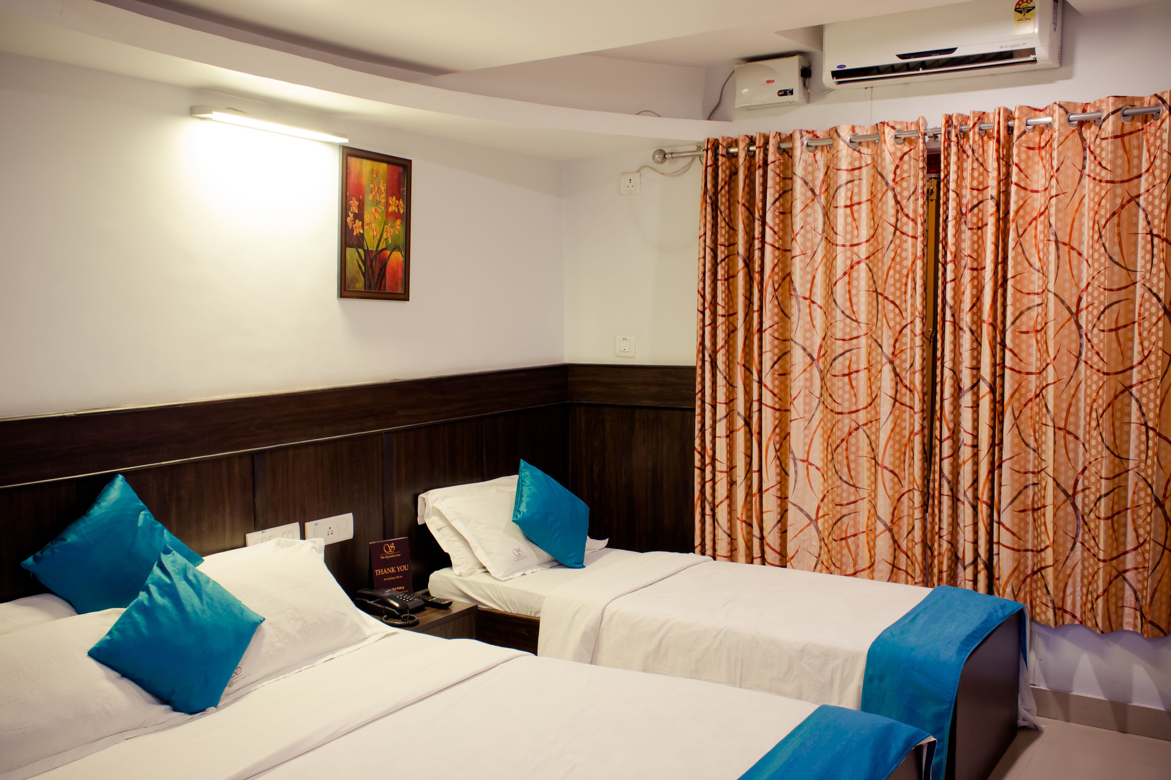 A hotel room with two beds, each with white bedding and blue pillows. There is a painting on the wall, a curtain with a leaf pattern, and an air conditioning unit above the beds.