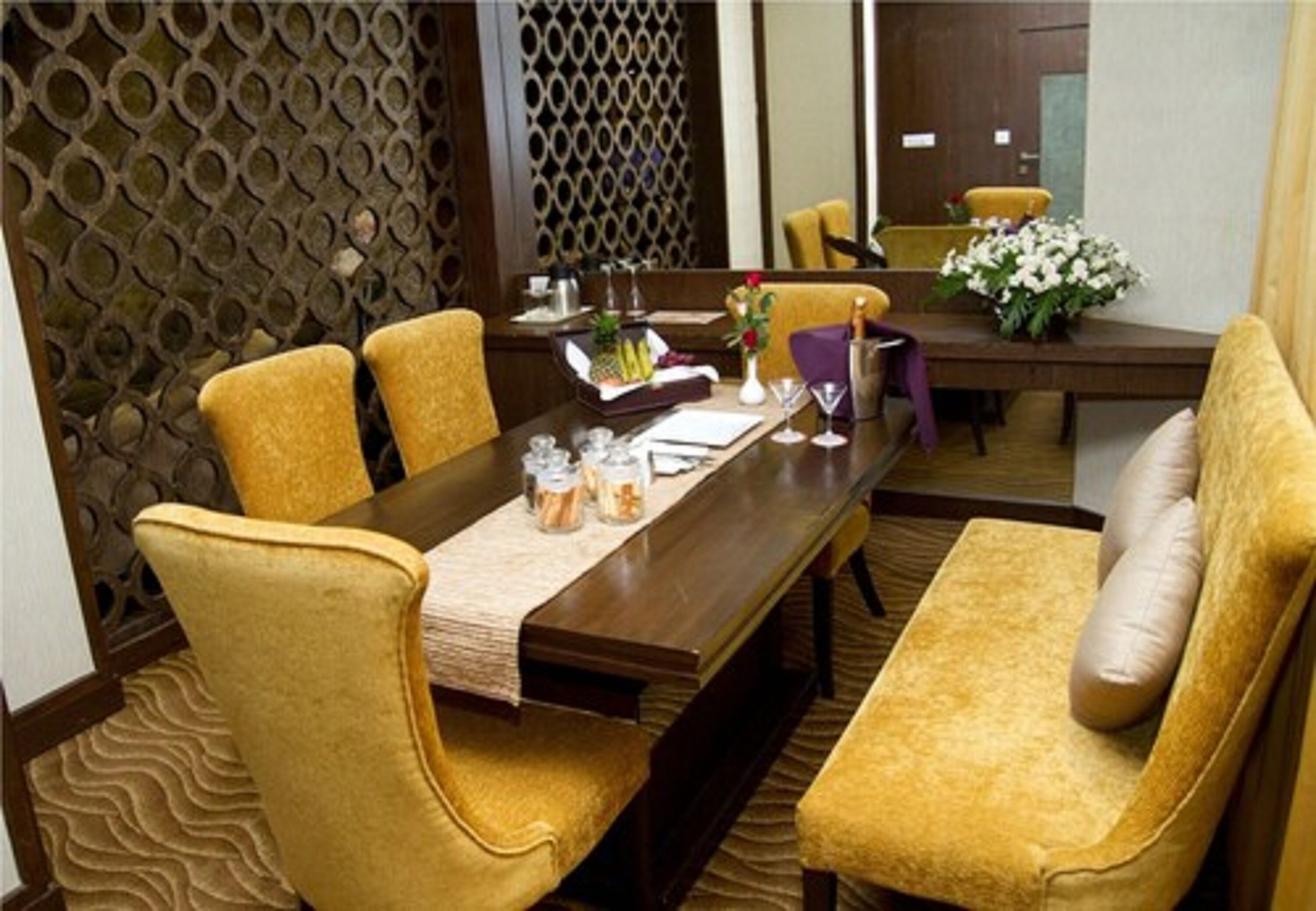 A dining area with a long wooden table, yellow upholstered chairs, and a decorative wall with a geometric pattern.