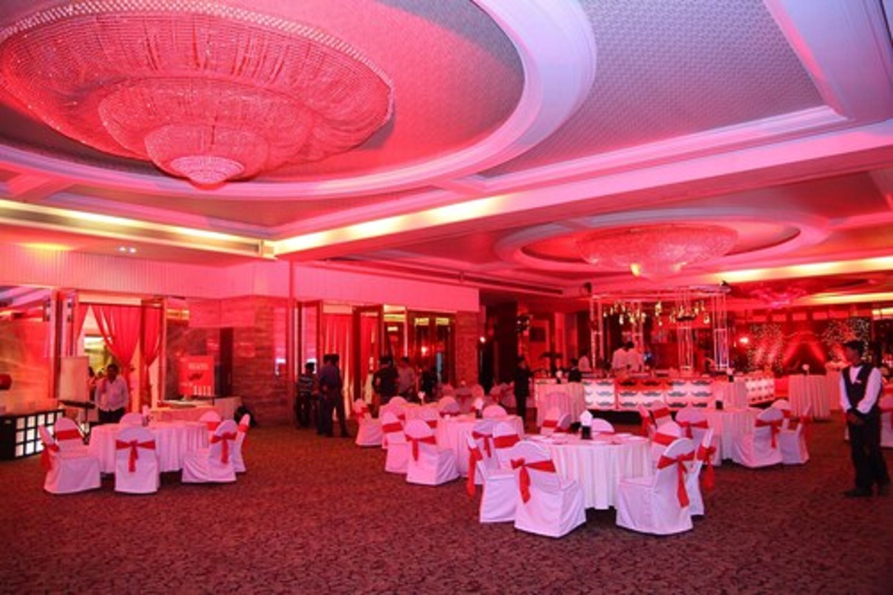 A banquet hall with round tables covered in white tablecloths and red sashes, a chandelier, and red ambient lighting.