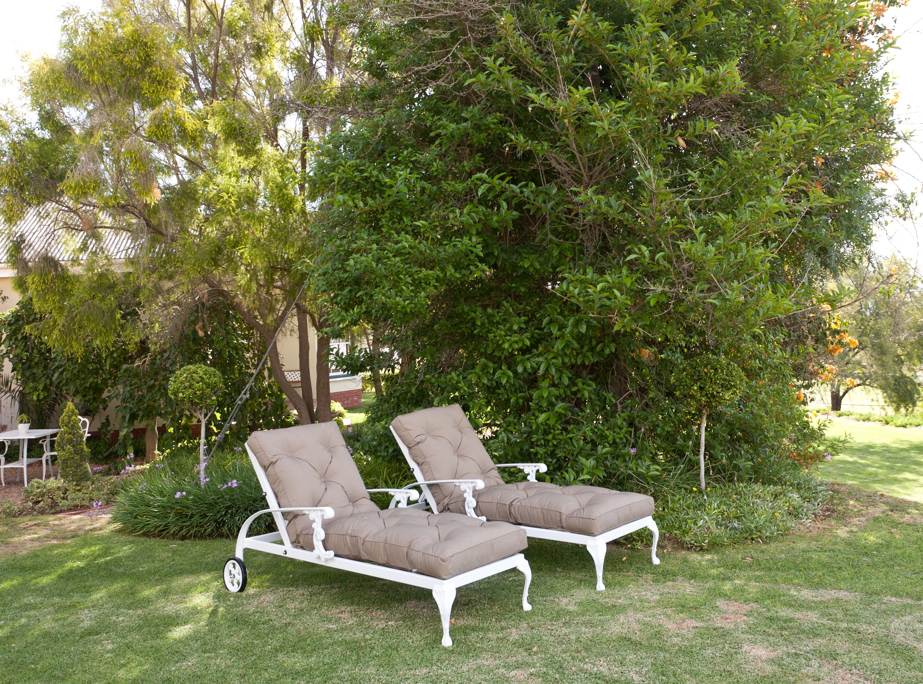 A white metal outdoor lounge chair with a cushioned seat and backrest, positioned on a grassy lawn with a large tree in the background.
