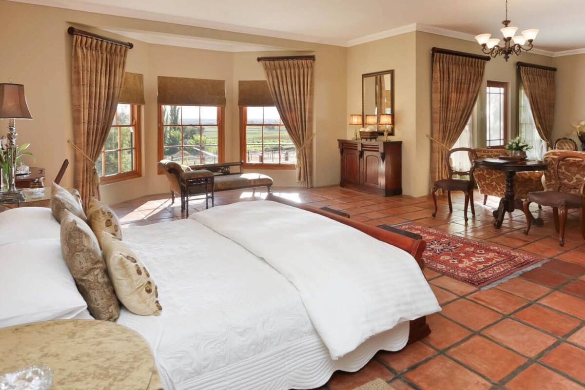 A spacious bedroom with a large bed, a desk, and a chair, along with a dining area and a view of the outdoors.