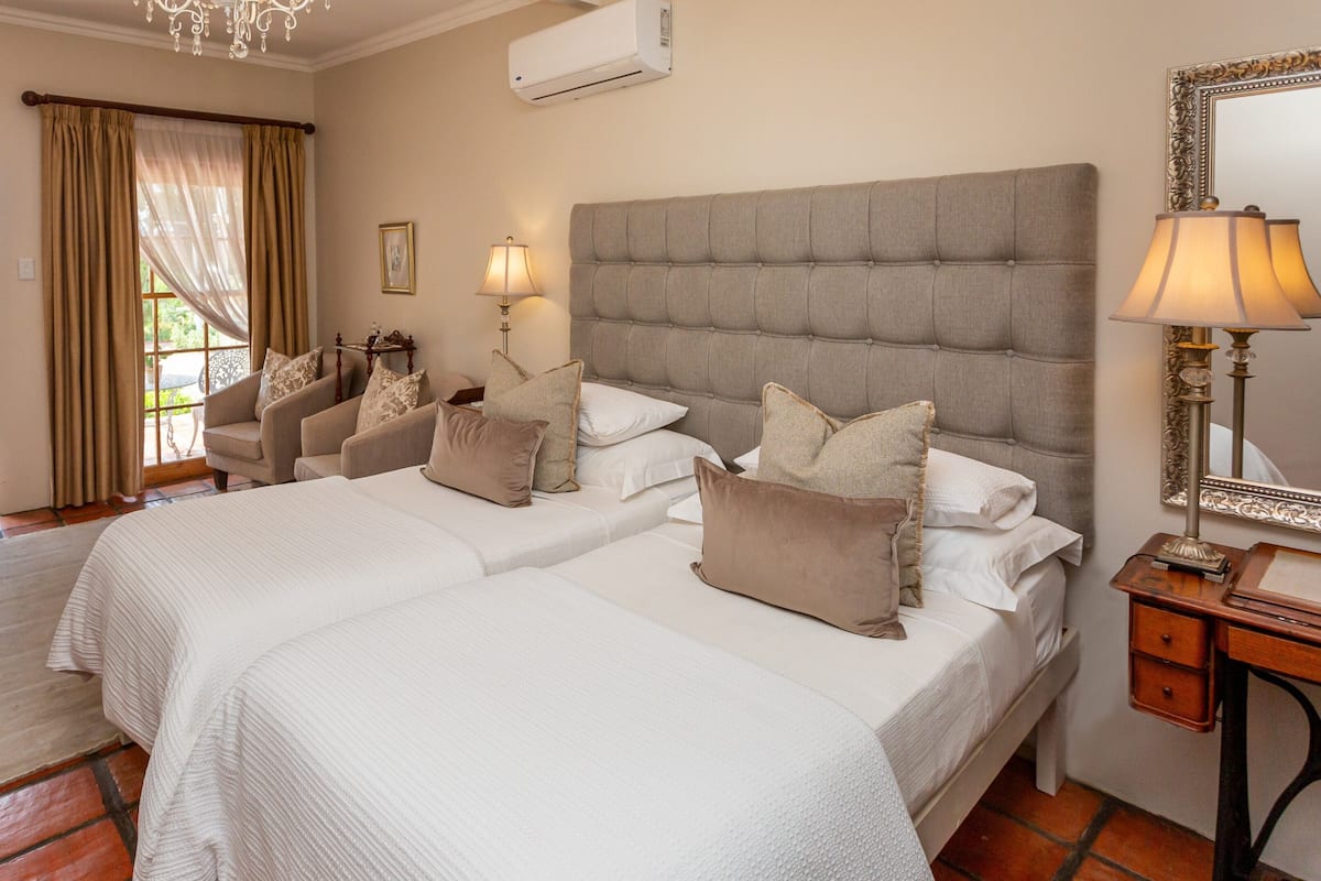 A hotel room with two beds, a large window with curtains, a chandelier, and a lamp on a bedside table.