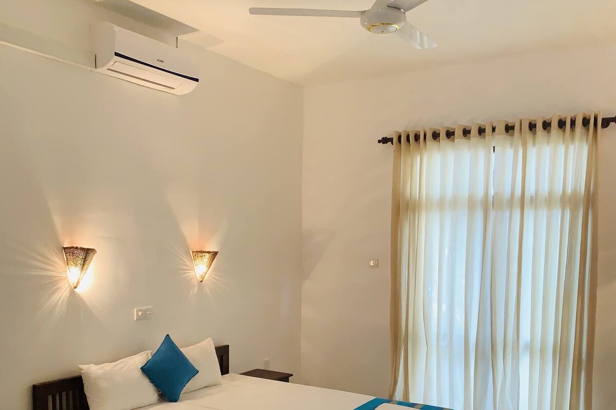 A hotel room with a bed, bedside tables, a ceiling fan, an air conditioning unit, and a window with curtains.