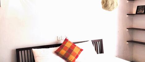 Un lit bien fait, agrémenté d’un coussin à carreaux, d’une suspension lumineuse et d’une étagère où sont posées des photos.