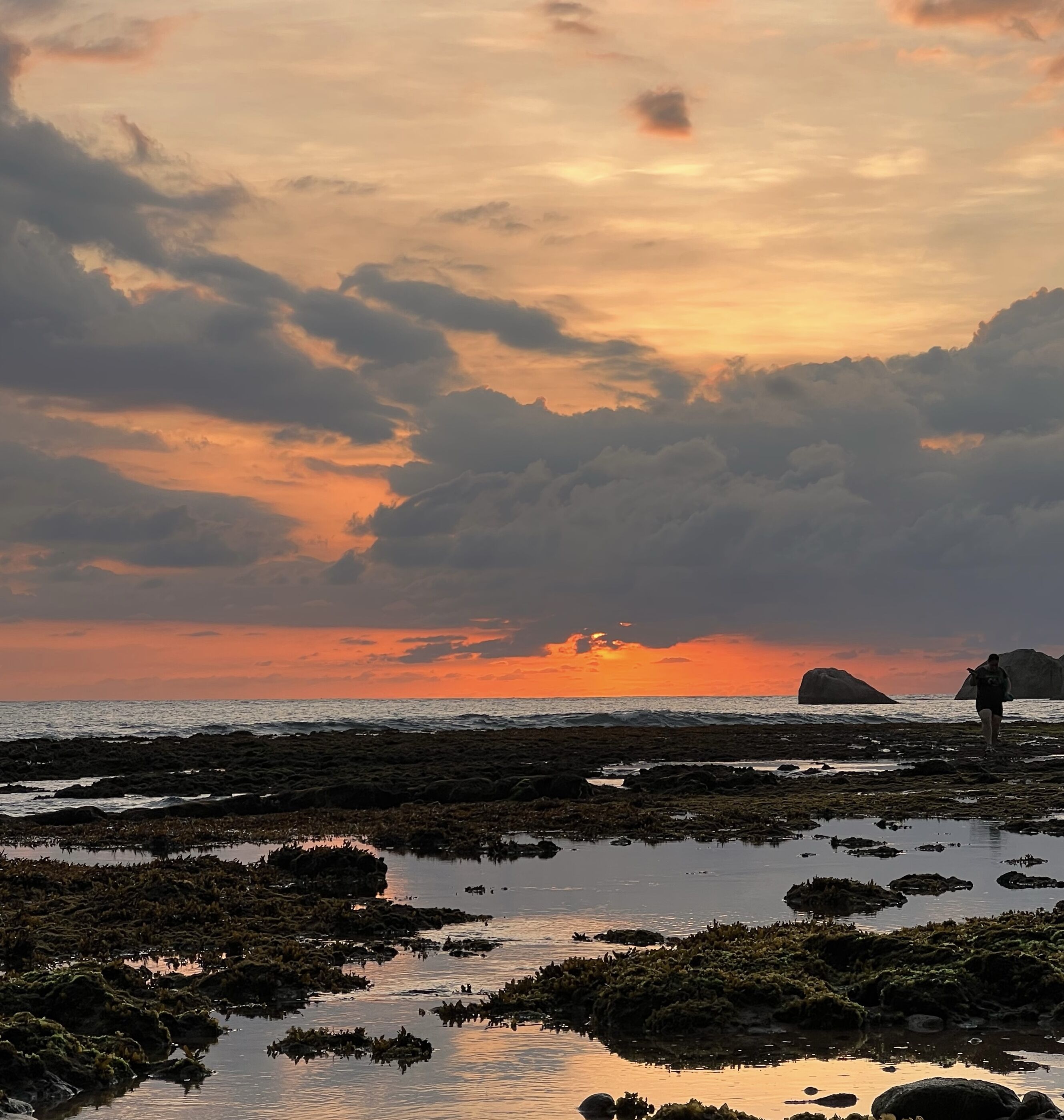 A rocky shoreline with a person carrying a surfboard, a large rock formation in the distance, and a sunset sky.