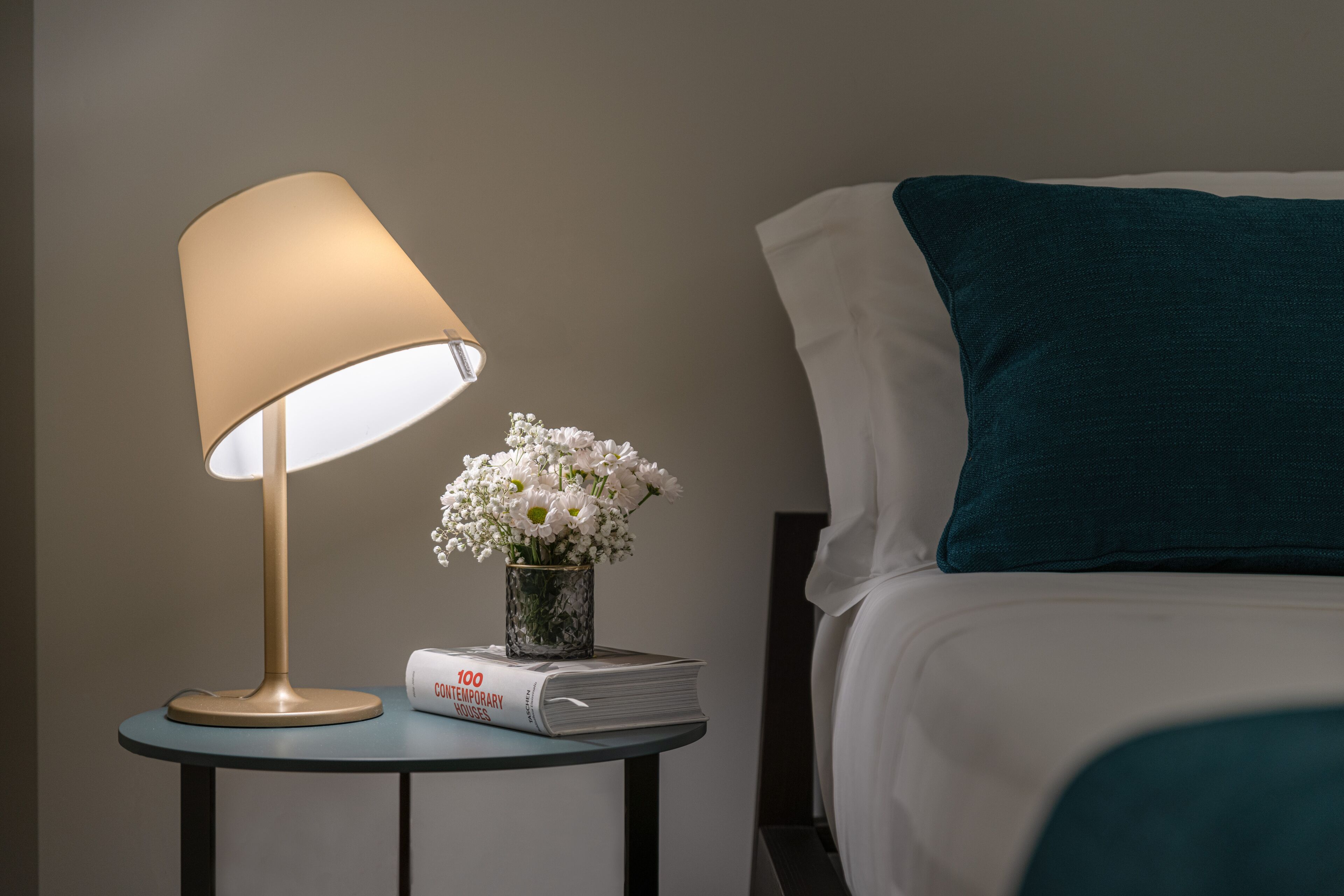 A neatly made bed with a teal pillow, a bedside lamp, a small vase with white flowers, and a book titled "100 Contemporary Houses".