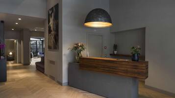 A modern reception desk with a wooden counter and a large pendant light. There is a vase with flowers on the counter and a mirror above it. The floor is made of herringbone-patterned wood.
