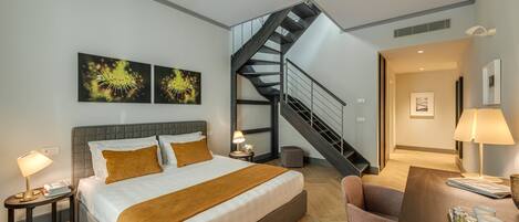 A modern hotel room with a large bed, a desk with a tray of items, a staircase, and artwork on the walls.