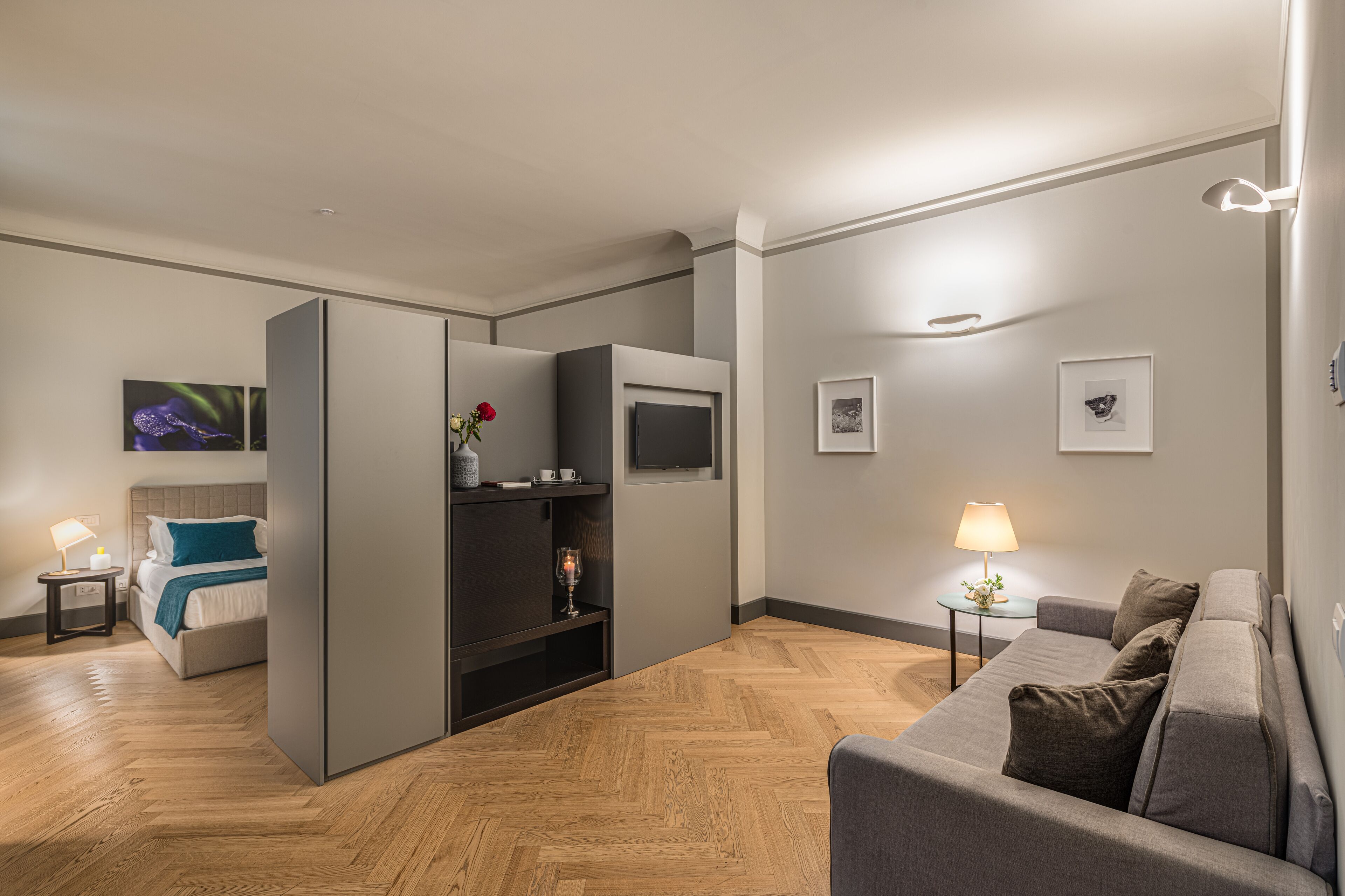 A modern hotel room with a bed, a sofa, a TV, and a small table with a lamp.