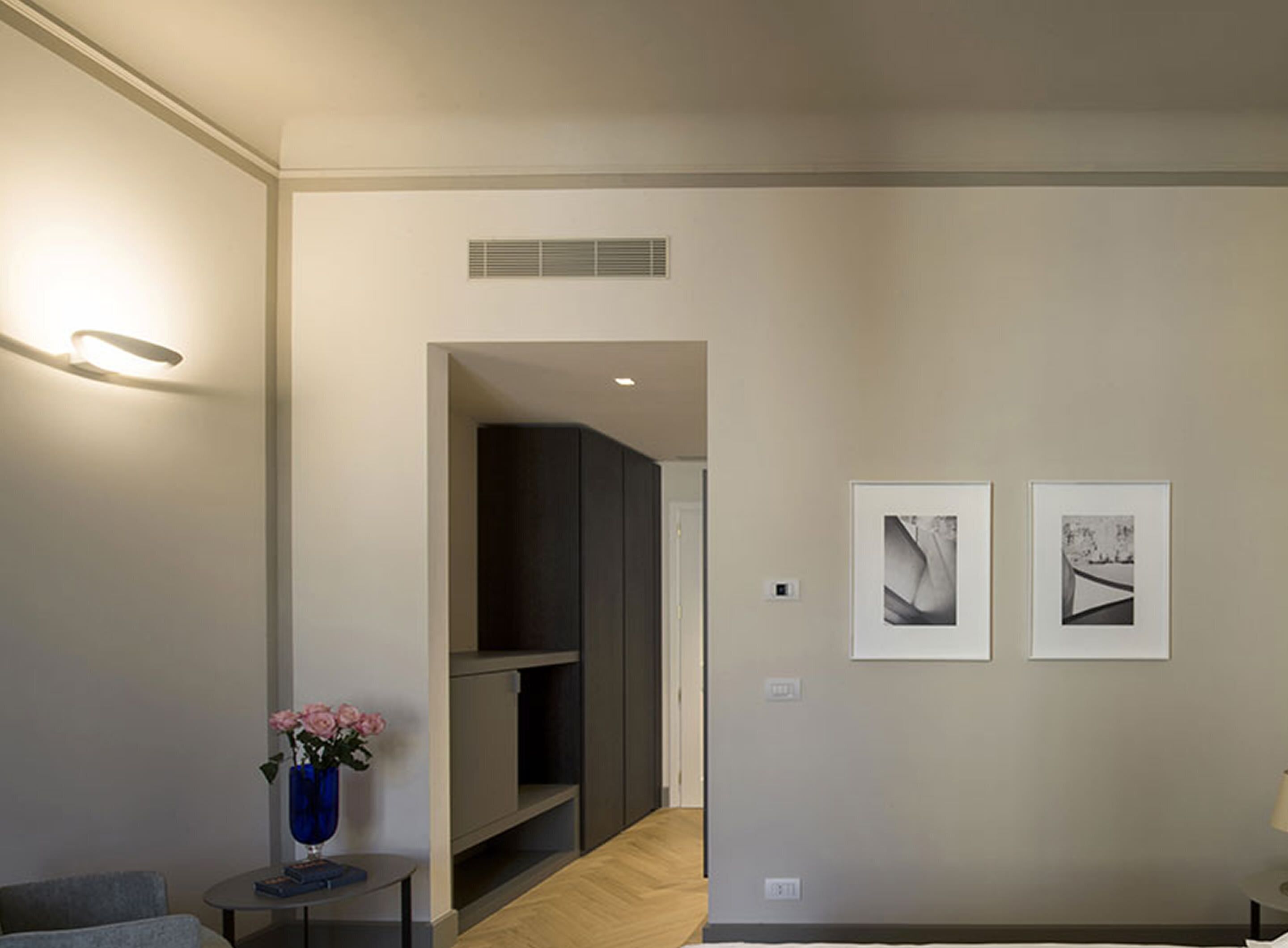 Una habitación de hotel moderna con un diseño minimalista, que presenta un mueble de madera oscura, una pequeña mesa con un jarrón de flores y dos obras de arte enmarcadas en la pared.