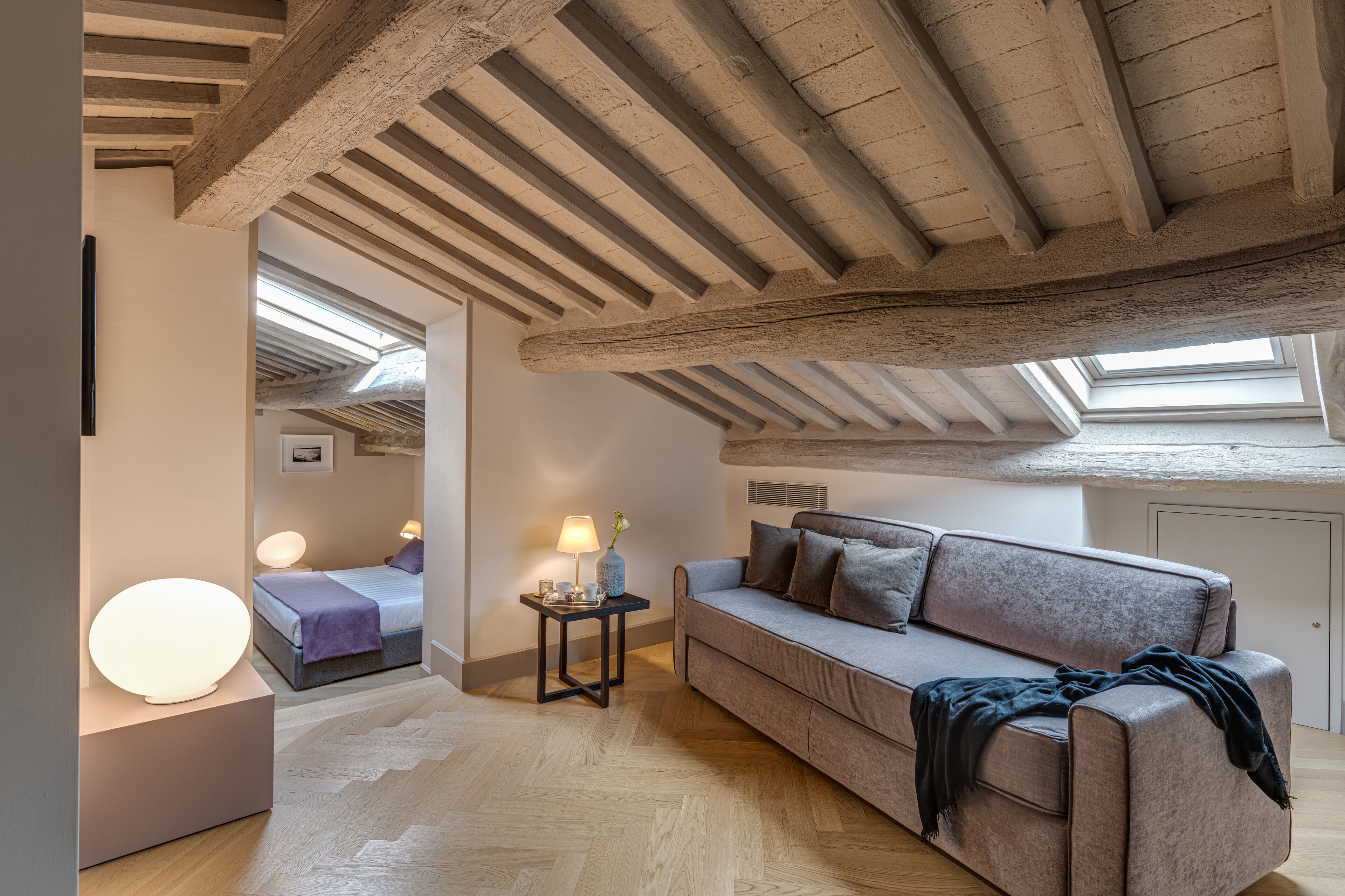 A modern loft bedroom with exposed wooden beams, a cozy gray sofa, a bedside table with a lamp, and a large mirror reflecting the room.