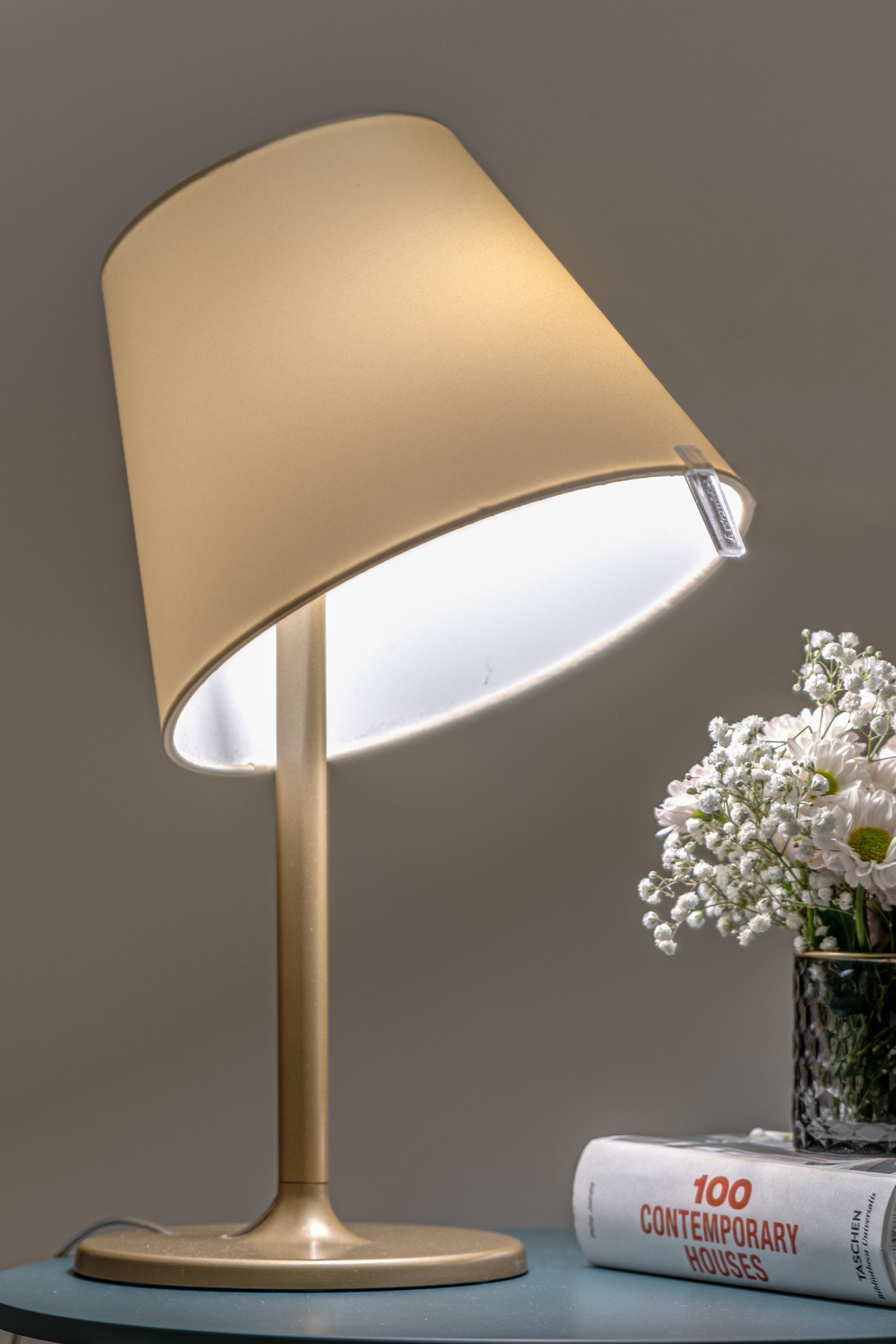 En lampa med en beige lampskärm, en bok med titeln ”100 moderna hus” och en vas med vita blommor på ett bord.
