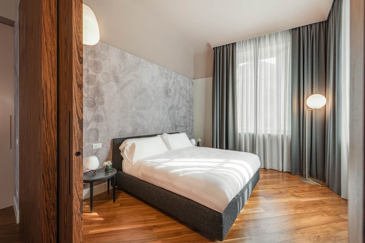 Habitación de hotel moderna con una cama grande, una mesita de noche, una lámpara de pared y vistas al exterior a través de cortinas translúcidas.