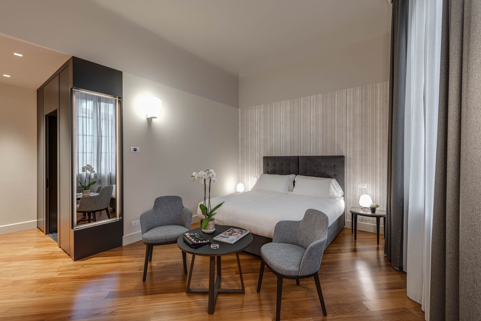 Una habitación de hotel moderna con una cama grande, dos sillones grises, una mesita redonda y un espejo.