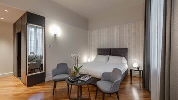 Una habitación de hotel moderna con una cama grande, dos sillones grises, una mesita redonda y un espejo.