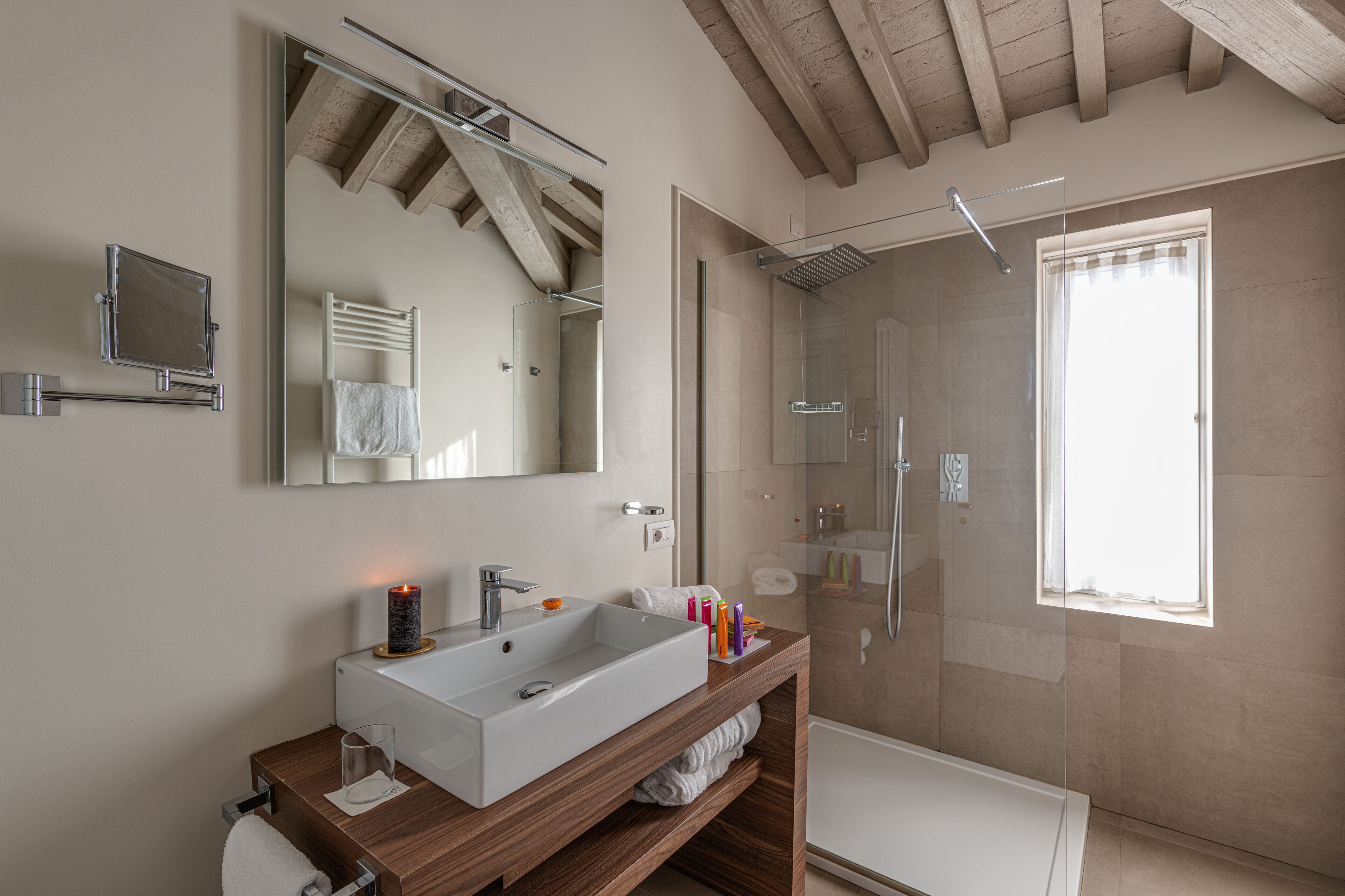 Ein modernes Badezimmer mit einem großen Spiegel, einem Holzanrichte, einem weißen Waschbecken, einer Duschkabine aus Glas und einem Fenster mit Tageslicht.