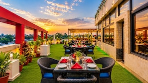 Een terras met tafels en stoelen, plantenbakken en uitzicht op de stad bij zonsondergang.