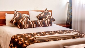 Een net opgemaakt bed met patroon kussens en een geweven deken, een houten hoofdbord en een witte muur met een stopcontact.
