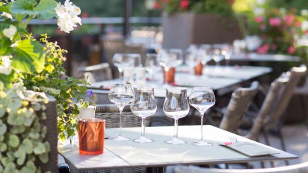 Une table à manger dressée avec des verres à vin et une bougie, entourée de chaises en osier et de plantes vertes.