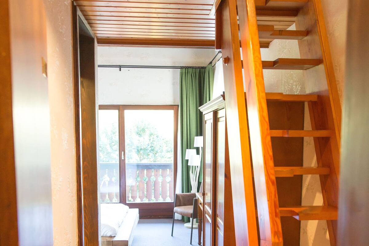 Un couloir étroit avec un escalier en bois menant à l’étage supérieur. On aperçoit un banc et une chaise à l’arrière-plan, près d’une grande fenêtre.