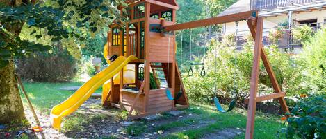 A wooden play structure with a slide and climbing area, surrounded by greenery and a house in the background.