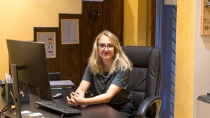 Une personne est assise à un bureau devant un ordinateur, avec une souris et un clavier. On aperçoit une chaise avec un repose-tête, une lampe de bureau et un certificat encadré accroché au mur.