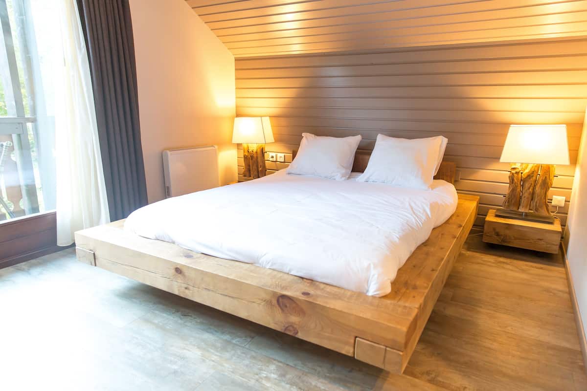 Une chambre moderne avec un lit surélevé en bois, deux tables de chevet avec des lampes et une vue sur l’extérieur.