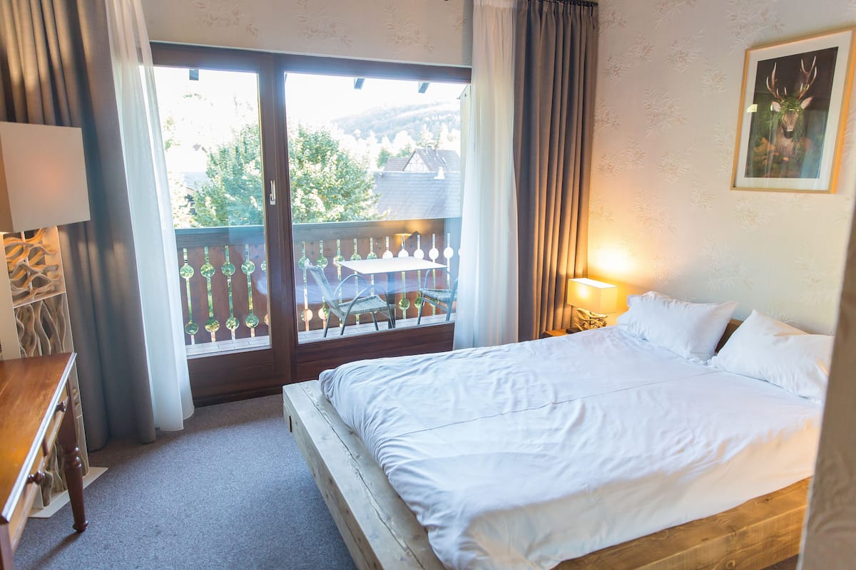 Une chambre d’hôtel avec un grand lit, une table de chevet en bois, une lampe, une fenêtre donnant sur des arbres et un balcon.