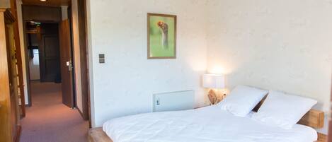 A bedroom with a wooden bed frame, white bedding, a nightstand with a lamp, and a framed picture on the wall.