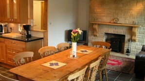 A dining area with a wooden table and chairs, a fireplace, and a kitchen in the background.