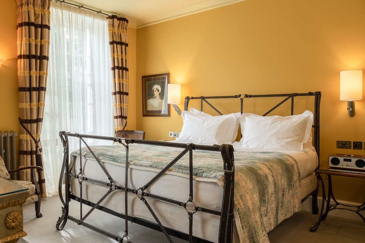A hotel room with a metal-framed bed, a chair, a table with a lamp, and a picture on the wall.